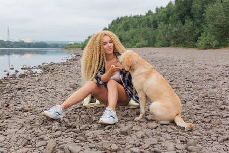 Young beautiful woman with blond curly hair sitting with her labrador retriever dog on the river shoreの写真素材