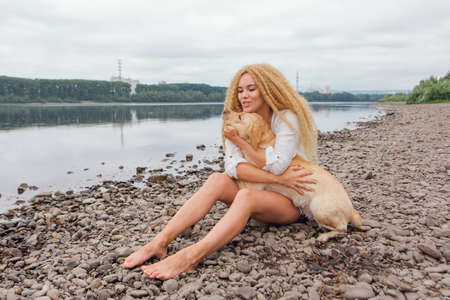 Young beautiful woman with blond curly hair sitting with her labrador retriever dog on the river shoreの写真素材