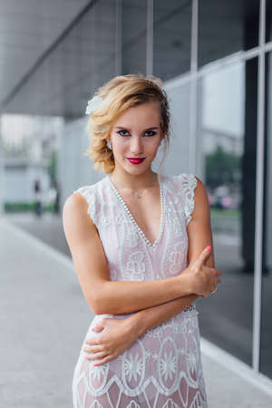 Young beautiful blond bride in white wedding dress and bright make up standing near mirror window outdoors.の写真素材