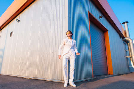 Wide angle portrait of a handsome man dressed in a white suit and round hippie sunglasses standing near the corner of a building during sunsetの写真素材