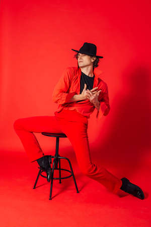 Portrait of a tall handsome man dressed in red shirt, red trousers and black hat posing with chair on the red backgroundの写真素材