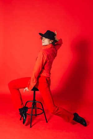 Portrait of a tall handsome man dressed in red shirt, red trousers and black hat posing with chair on the red backgroundの写真素材