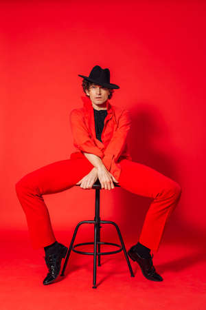 Portrait of a tall handsome man dressed in red shirt, red trousers and black hat posing with chair on the red backgroundの写真素材