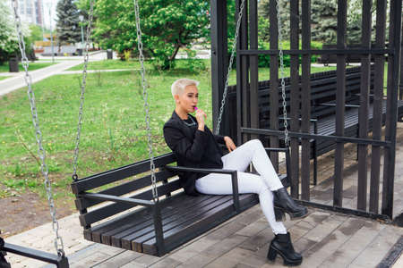 Young stylish business woman with short hair and nose piercing. Confident girl look like lesbian sitting and smoking electronic cigarette on the swingの写真素材