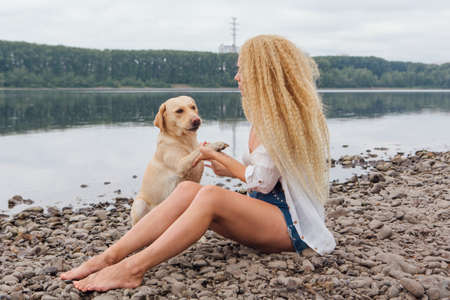 Young beautiful woman with blond curly hair sitting with her labrador retriever dog on the river shoreの写真素材