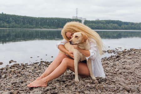 Young beautiful woman with blond curly hair sitting with her labrador retriever dog on the river shoreの写真素材