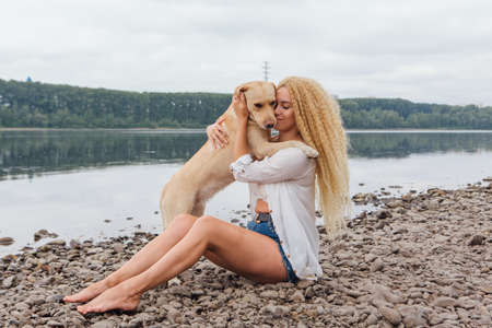 Young beautiful woman with blond curly hair sitting with her labrador retriever dog on the river shoreの写真素材
