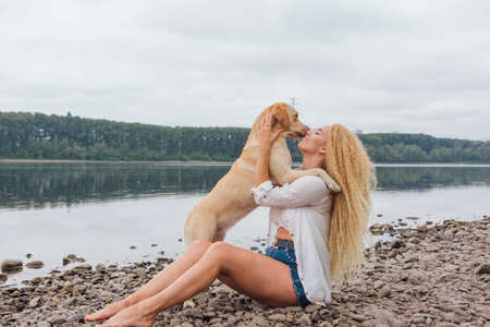 Young beautiful woman with blond curly hair sitting with her labrador retriever dog on the river shoreの写真素材