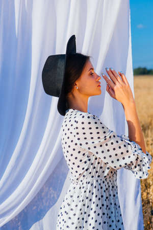 Rural Countryside Scene. Young beautiful woman with long hair dressed in white Polka-dot dress and black hat standing at golden oat field with a white curtain on the background.の写真素材
