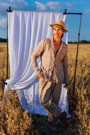Rural Countryside. Tall handsome man dressed in a coarse linen suit and hat standing at golden oat field with a white curtain on the background.の写真素材