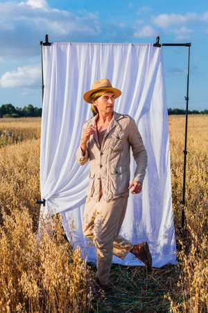 Rural Countryside. Tall handsome man dressed in a coarse linen suit and hat standing at golden oat field with a white curtain on the background.の写真素材