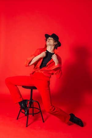 Portrait of a tall handsome man dressed in red shirt, red trousers and black hat posing with chair on the red backgroundの写真素材