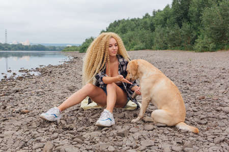 Young beautiful woman with blond curly hair sitting with her labrador retriever dog on the river shoreの写真素材