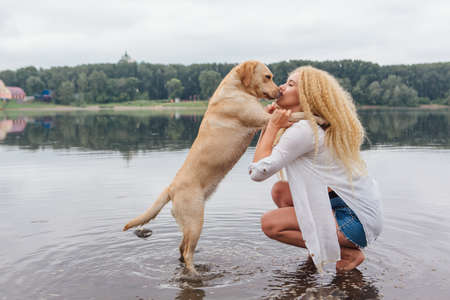Young beautiful woman with blond curly hair huging with her labrador retriever dog on the river shoreの写真素材
