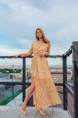 Beautiful young woman in long leopard dress standing on the stairs on the top of the roof of a building.の写真素材