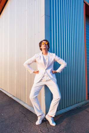 Wide angle portrait of a handsome man dressed in a white suit and round hippie sunglasses standing near the corner of a building during sunsetの写真素材