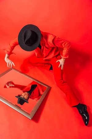 Tall handsome man dressed in red shirt and black hat sitting on the red background and looking into the mirror on his own reflection.の写真素材