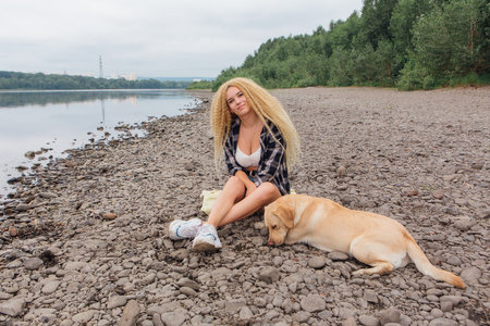 Young beautiful woman with blond curly hair sitting with her labrador retriever dog on the river shoreの写真素材
