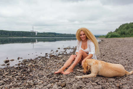 Young beautiful woman with blond curly hair sitting with her labrador retriever dog on the river shoreの写真素材