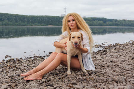 Young beautiful woman with blond curly hair sitting with her labrador retriever dog on the river shoreの写真素材