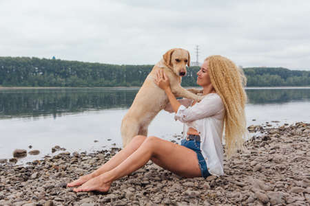 Young beautiful woman with blond curly hair sitting with her labrador retriever dog on the river shoreの写真素材