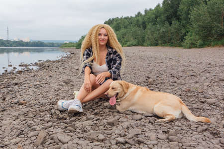 Young beautiful woman with blond curly hair sitting with her labrador retriever dog on the river shoreの写真素材