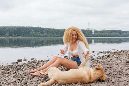 Young beautiful woman with blond curly hair sitting with her labrador retriever dog on the river shoreの写真素材