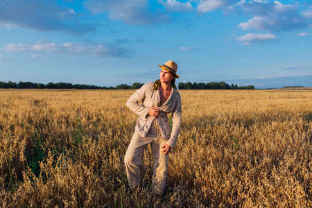 Rural Countryside. Tall handsome man dressed in a coarse linen suit and hat standing at golden oat fieldの写真素材