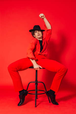 Portrait of a tall handsome man dressed in red shirt, red trousers and black hat posing with chair on the red backgroundの写真素材