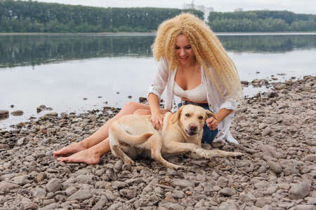 Young woman sitting with her labrador retriever dog on the river shoreの写真素材