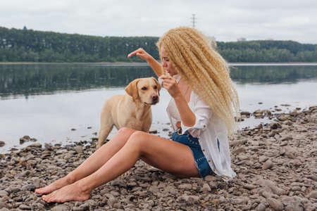 Young beautiful woman with blond curly hair sitting with her labrador retriever dog on the river shoreの写真素材