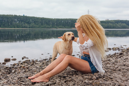 Young woman sitting with her labrador retriever dog on the river shoreの写真素材
