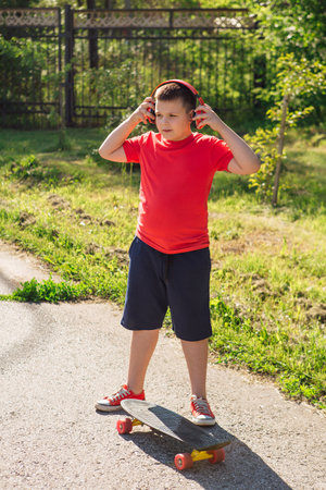 Happy chubby ten years old boy with headphones outdoors. Boy listening to music. Teen using phone at summer park. Fashion, lifestyle, technologyの写真素材