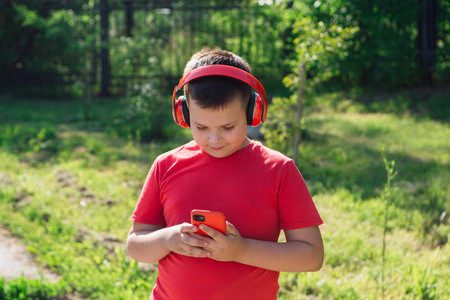 Happy chubby ten years old boy with headphones outdoors. Boy listening to music. Teen using phone at summer park. Fashion, lifestyle, technologyの写真素材