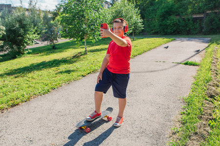 Chubby ten years old boy spends free time training skating in the city park. boy looking on mobile phone. Cute nice boy enjoys sunny day outdoors with skateboard.の写真素材