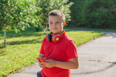 Happy chubby ten years old boy with headphones outdoors. Boy chatting on mobile. Teen using phone at summer park. Fashion, lifestyle, technologyの写真素材