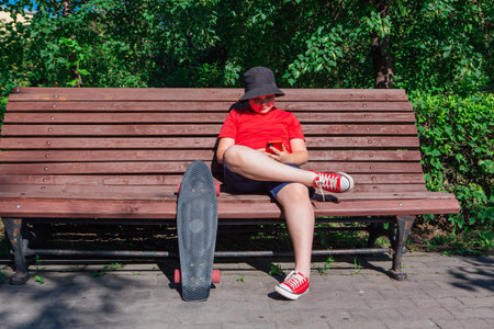 Boy with plastic skateboard sitting on the bench and stares at the phone.の写真素材