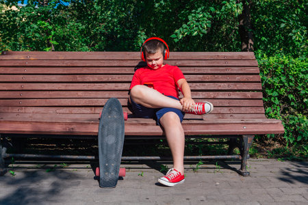 Boy with plastic skateboard sitting on the bench, stares at the phone and listening music in headphones.の写真素材
