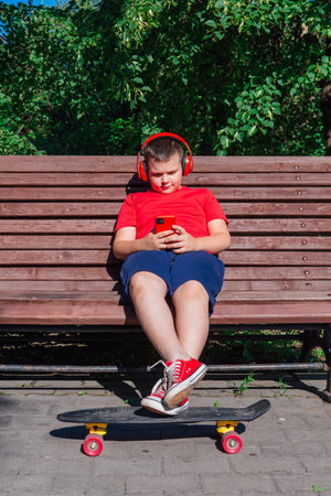 Boy with plastic skateboard sitting on the bench, stares at the phone and listening music in headphones.の写真素材