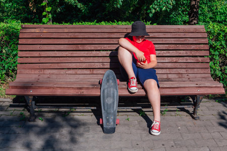 Boy with plastic skateboard sitting on the bench and stares at the phone at hot summer day.の写真素材