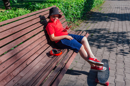 Boy with plastic skateboard sitting on the bench, stares at the phone at hot summer day.の写真素材