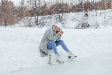 Lovely young woman relaxing after riding ice skates and drinking hot drink from thermo pot on the ice rink. Girl sitting on snow and smiling.の写真素材