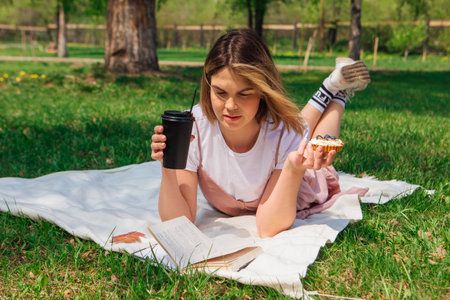 Young woman laying on the grass in park with open book, cake and cup of coffee in a sunny summer dayの写真素材