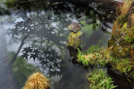 Old stone doll in ancient pool with reflection of Japanese maple  and rocks with trees up poolside...