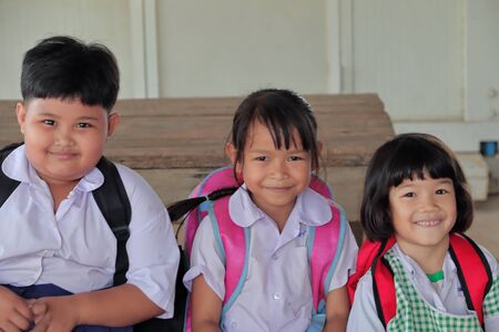 3 lovely girls in rural area wearing school uniform, school bag.   Sit on the wooden chair and smiling with joy to photograph.のeditorial素材