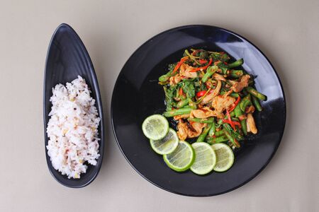 Thai cuisine , Spicy fried cowpea and basil with pork topped sliced green lemon served with japanese seed for rice in jasmine rice on black platesの写真素材
