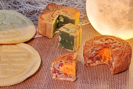 Round mooncake filled 8 grains and salted egg  and square mooncake filled red bean paste stirred in green tea and chopped macadamia nuts served with  two green bean mooncakes placed on sackcloth near moon shaped light.の写真素材