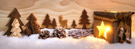 Beautiful Christmas arrangement with nice wooden ornaments and shining lantern, with wood board background, illustrating a conifer forest in snow, extra wide formatの写真素材