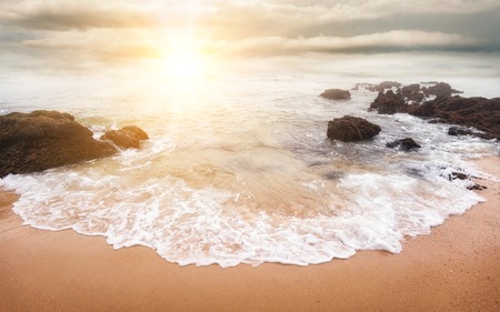 Glorious sunrise scene at the ocean with the sun, clouds and a beautiful little white wave on the sand in the foregroundの写真素材