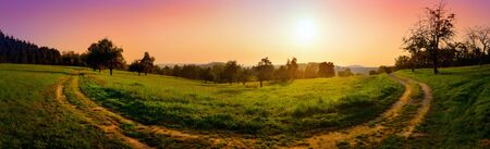Dreamy and dramatic sunrise over rural landscape: colorful panorama shot of a curved path on a meadow with red and purple skyの写真素材
